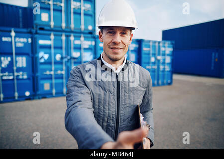 Port manager wearing a hardhat extending a handshake while standing on a large commercial shipping dock holding a clipboard Stock Photo