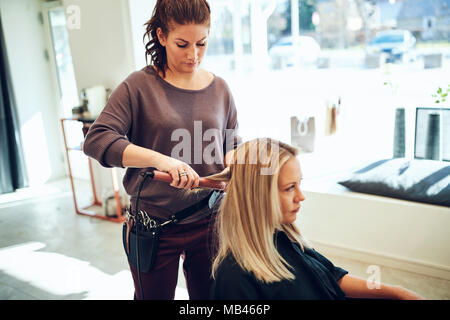 Young stylist styling the hair a female client sitting in a chair with a straightener while working in her salon Stock Photo