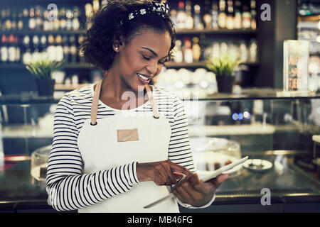 Smiling African entrepreneur wearing an apron using a digital tablet while standing in front of a counter of her trendy cafe Stock Photo