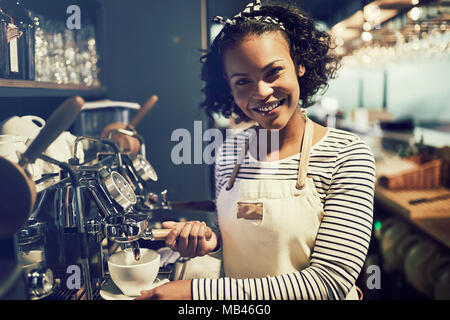 Young African barista smiling and preparing a fresh cup of coffee while standing at a coffee maker in a trendy cafe Stock Photo