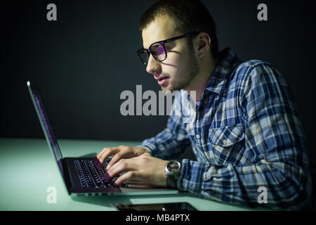 Concentrated young hacker using computer and stealing information in dark room Stock Photo