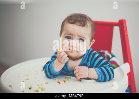 6 month old baby boy feeding himself in a high chair Stock Photo