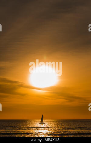 Lone sailboat sailing on a calm ocean under a vivid orange tropical sunset silhouetted against the reflection cast by the sun over the water Stock Photo