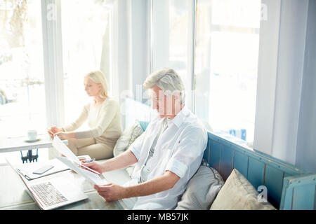 Relaxed mature businessman reading financial papers while sitting in cafe after lunch with woman on background Stock Photo