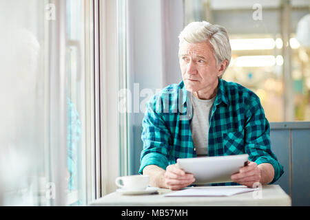 Pensive senior man with tablet looking through window of cafe while thinking of working points Stock Photo