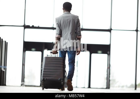 Rear view of young modern traveler pulling suitcase after himself while walking towards exit to flight station Stock Photo