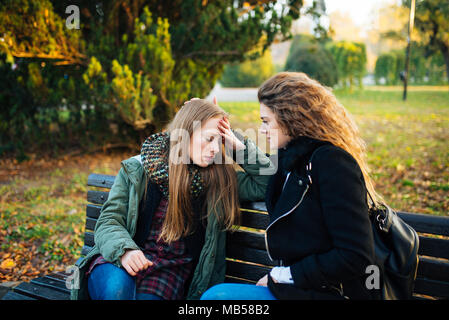 Sad attractive girl sitting on the bench in the park while her female friend is consoling her. Stock Photo