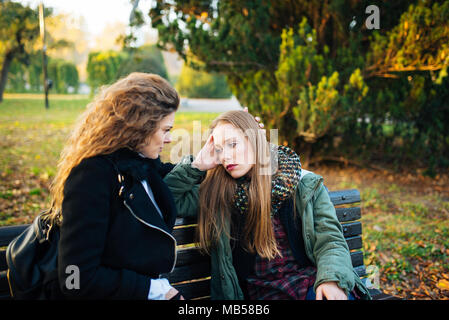 Sad attractive girl sitting on the bench in the park while her female friend is consoling her. Stock Photo