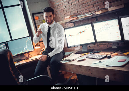 Young bearded man trader at office sitting on desk drinking hot coffee morning refreshment smiling cheerful Stock Photo