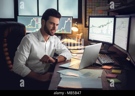 Young bearded man trader at office sitting at table using laptop looking at screen smiling satisfied good performance Stock Photo