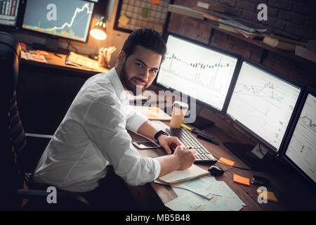 Young bearded man trader at office sitting at table looking camera smiling happy working Stock Photo