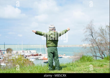 one little boy in jacket and hat stands on green hill on background of sea Bay with ships in early spring Stock Photo