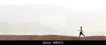 Young boy in orange t shirt walking in desert next to rope fence with misty mountains behind, Striding forth. Walking Stock Photo