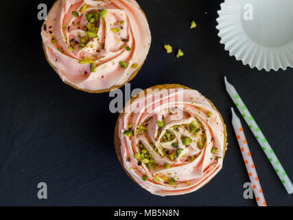 Two pink cup cakes with pistachio nut sprinkles and candles on black background, top view photograph Stock Photo
