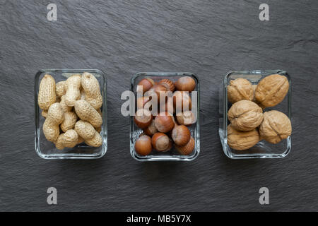 Peanuts, Hazelnuts and Walnuts with nutshells placed in glass plates on black stone background surface with free space Stock Photo
