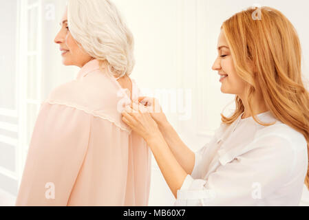 Mindful mature daughter assisting her senior mom in dressing up Stock Photo