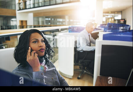 Pretty dreamy African-American office working woman sitting and talking phone at workplace. Stock Photo