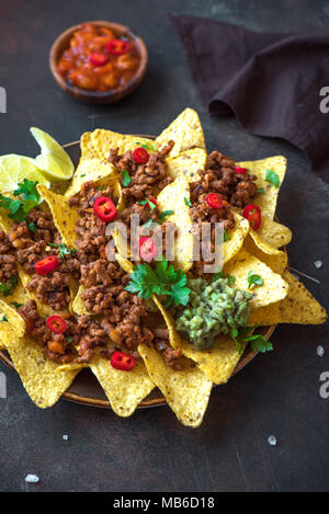 Mexican nacho corn tortilla chips with cheese, meat, guacamole and red hot spicy salsa. Nachos with ground beef on dark rustic background, copy space. Stock Photo