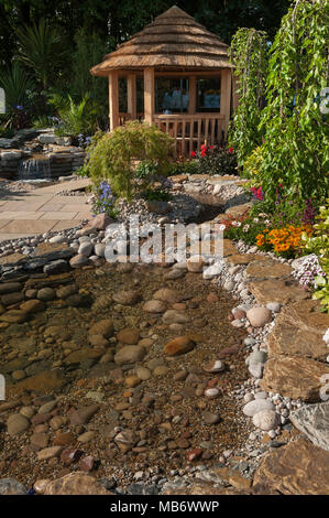 Beautiful spot to rest (summerhouse, seating, stream & bright flowers) - 'Water Way to Go ' rock garden, RHS Flower Show, Tatton Park,  England, UK. Stock Photo