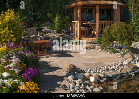 Beautiful spot to rest (summerhouse, seating, stream & bright flowers) - 'Water Way to Go ' rock garden, RHS Flower Show, Tatton Park,  England, UK. Stock Photo