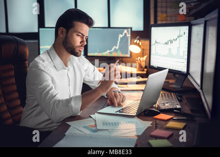 Young bearded man trader at office sitting at table looking at laptop thoughtful strategy planning Stock Photo