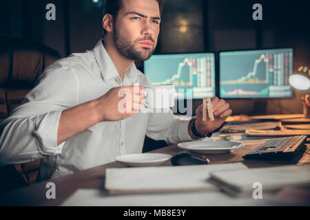 Young bearded man trader at office sitting at table drinking hot coffee eating sandwich working at night energy restoring Stock Photo