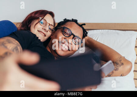 Cheerful multiracial couple lying together in bed and taking selfie. Smiling man and woman making selfie with mobile phone. Stock Photo