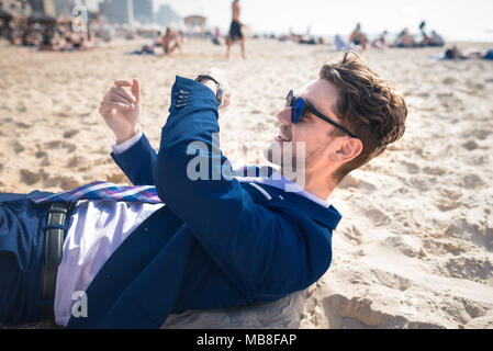Attractive young man in elegant suit has time to rest. Funny guy relaxes on the beach and looks on the wristwatch Stock Photo