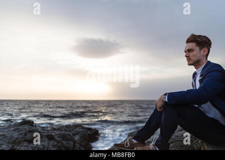 Serious young man in elegant suit sits on the coast near ocean. Young guy thinking on background of the water and sky Stock Photo