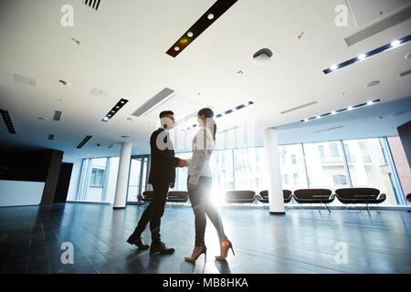 Smiling young entrepreneur wearing classical suit shaking hand of his business partner while greeting her at modern office lobby, lens flare Stock Photo
