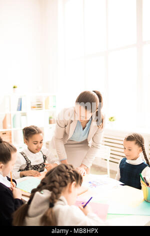 Teacher teaching kids in classroom at school. They are sitting together at the table and drawing Stock Photo