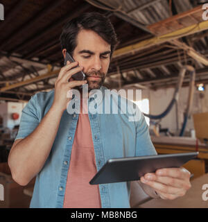 Young woodworker using a phone and tablet in his workshop Stock Photo