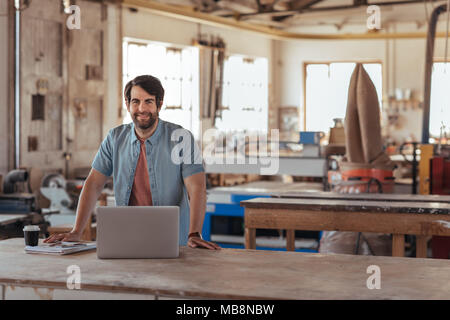 Smiling woodworker creating designs on a laptop in his workshop Stock Photo