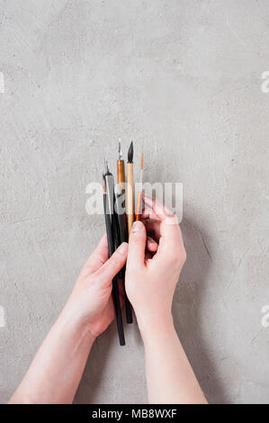 Woman’s hands holding the art brushes and ink pens on the textured grey table, top view Stock Photo