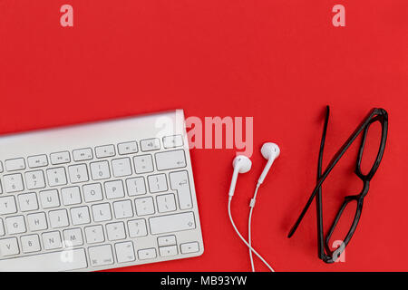 Computer keyboard white music earphones + eyeglasses on bright red background Stock Photo