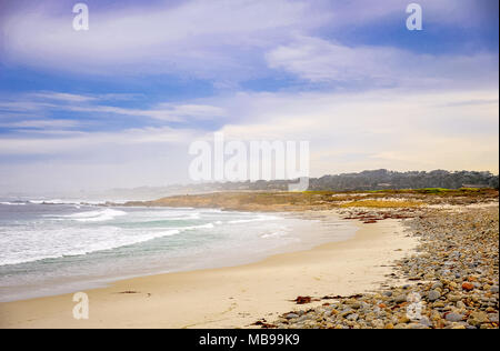 Pacific ocean seascape at Pebble beach near Monterey, California. Foggy background, crashing waves, sandy shore, colourful rocks and blue, cloudy sky Stock Photo