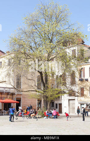 Campo San Polo, San Polo, Venice, Veneto, Italy in spring with locals and tourists relaxing on colorful red benches under a tree Stock Photo