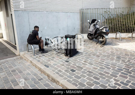 Paris, France - August 22, 2016: A Deliveroo delivery man is waiting for a call seated near his bike and food delivery bag laying on the ground. Stock Photo