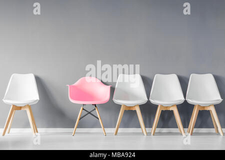 Front view of a row of modern, white chairs and a pink one against gray wall in a minimal style interior Stock Photo