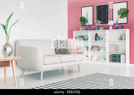 White sofa in pink living room interior with white brick wall, striped rug, posters and bookcase with books and decorations Stock Photo