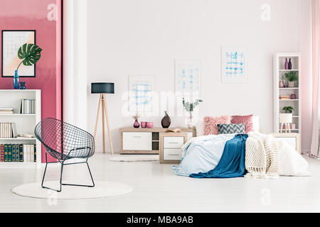 Big bed with cushions and sheets in modern bedroom interior with metal chair, round rug, posters on white wall, bookcases and black lamp Stock Photo