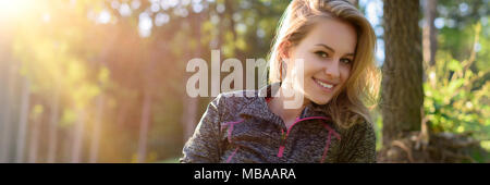 Attractive smiling young blond woman, wearing sportswear, relaxing after workout in a forest. Web banner. Stock Photo