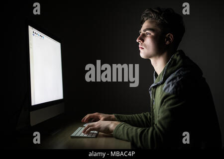 Concentrated young man in glasses sitting at the table and using computer in dark room Stock Photo