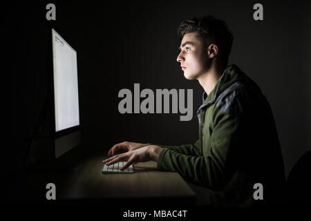 Concentrated young man in glasses sitting at the table and using computer in dark room Stock Photo