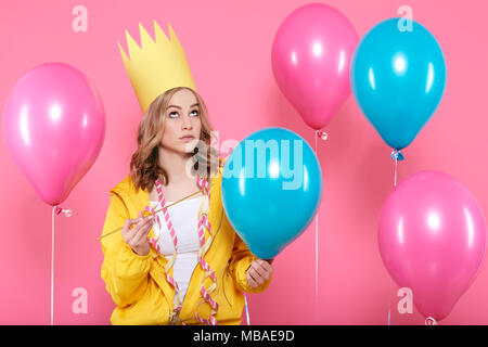 Funny conceptual photography. Cheeky girl in birthday hat holding needle pretending to pop birthday balloons. Attractive trendy teenager celebrating. Stock Photo