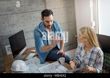 Young worried architect colleagues talking and analyzing blueprints while working on a new project on digital tablet at the office. Stock Photo