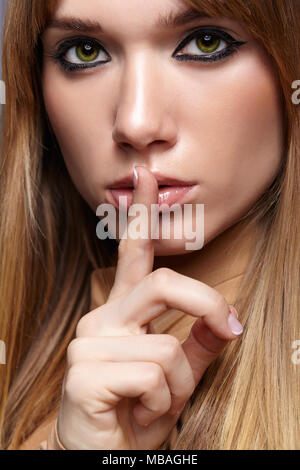 Portrait of blonde young woman with finger on lips. Female with green eyes and long straight hair. Girl dressed in beige dress on gray background. Stock Photo