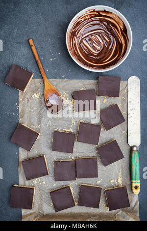 Caramel shortbread / Millionaires shortbread squares with a pallet knife and melted dark chocolate in a bowl on a slate background Stock Photo