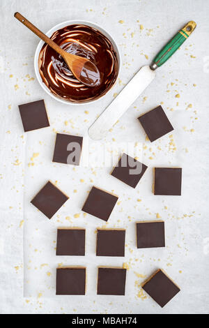 Caramel shortbread / Millionaires shortbread squares with a pallet knife and melted dark chocolate in a bowl on a white background Stock Photo