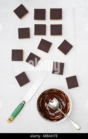 Caramel shortbread / Millionaires shortbread squares with a pallet knife and melted dark chocolate in a bowl on a white background Stock Photo